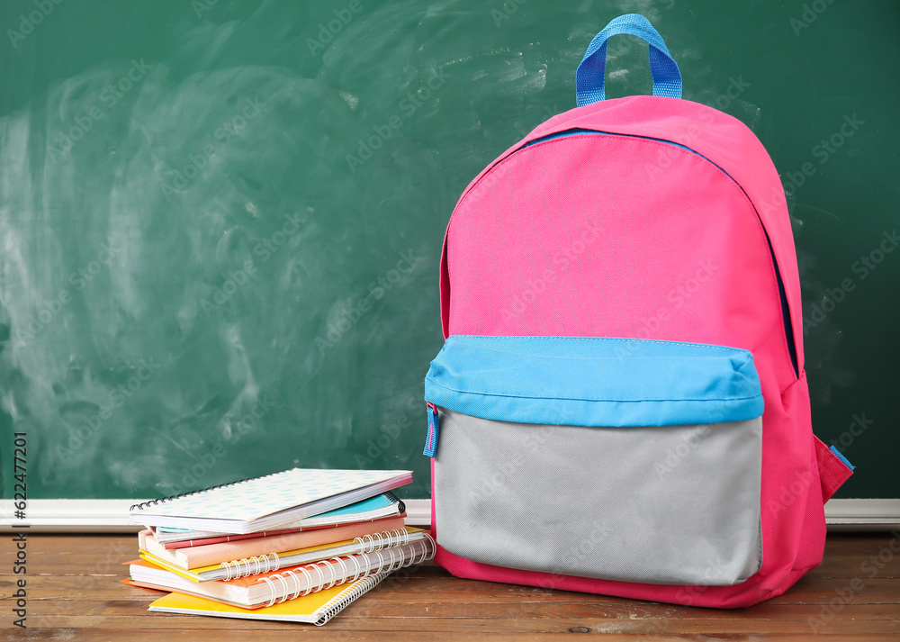 Pink school backpack with notebooks on wooden table near green chalkboard