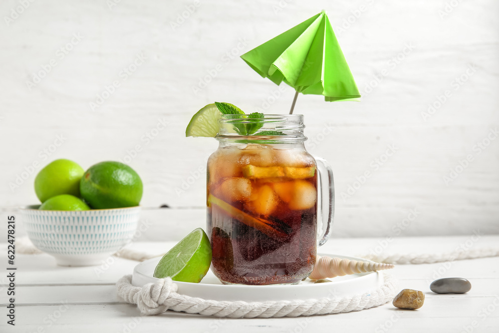 Mason jar of cold Cuba Libre cocktail and bowl with limes on white wooden