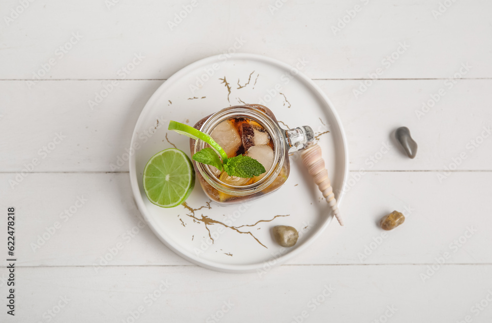 Plate with mason jar of cold Cuba Libre cocktail, stones and seashell on white wooden background