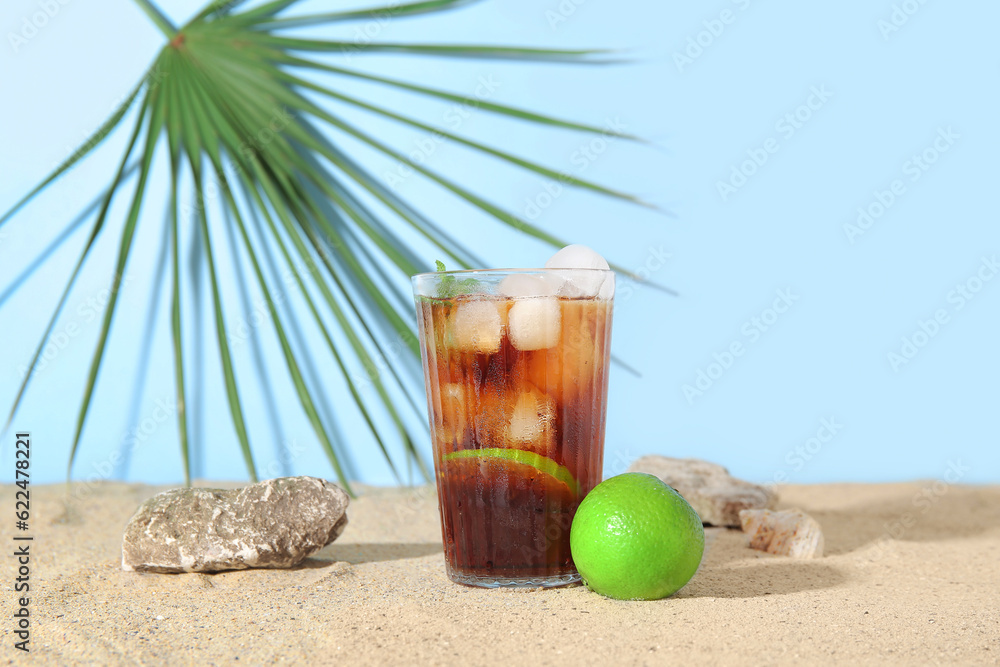 Glass of cold Cuba Libre cocktail, stones and palm leaf on sand near blue background