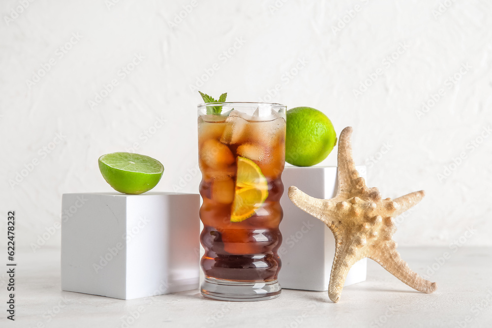 Glass of cold Cuba Libre cocktail, decorative podiums with limes and starfish on white background