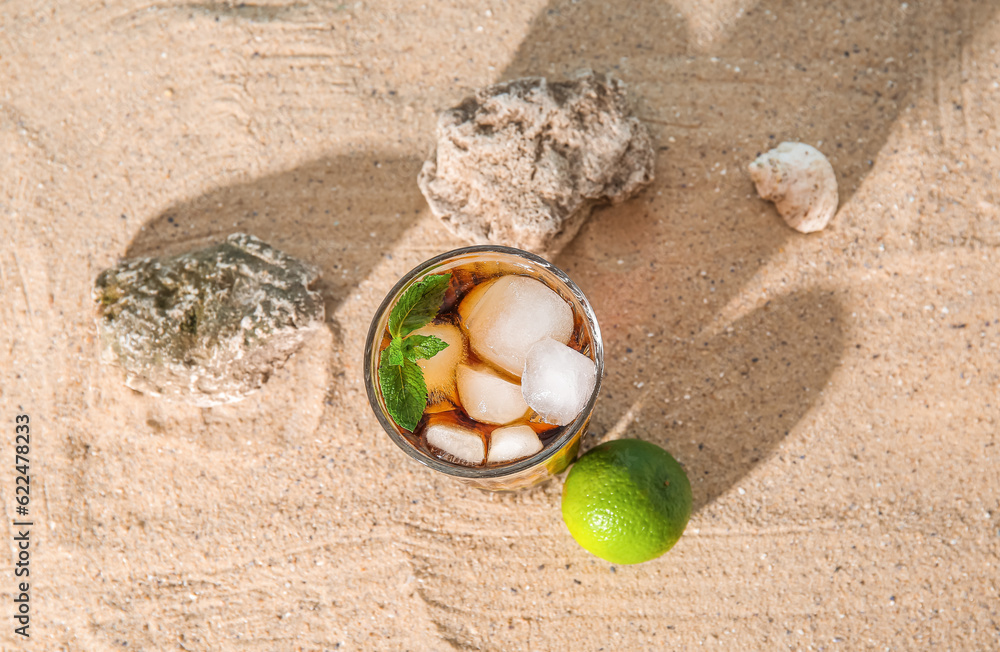 Glass of cold Cuba Libre cocktail and stones on sand