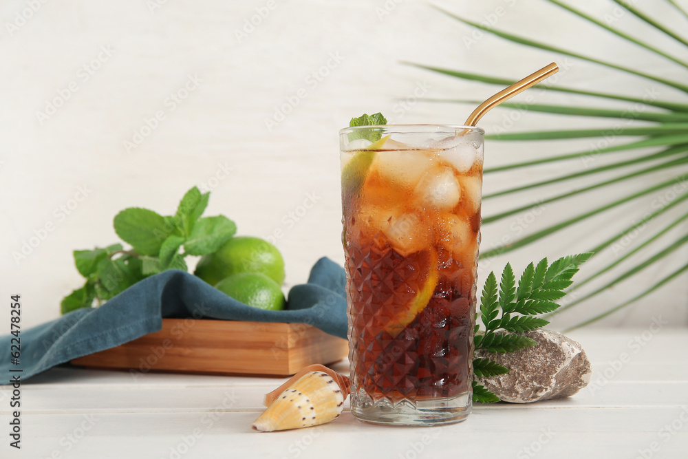 Glass of cold Cuba Libre cocktail, stone and seashells on white wooden table