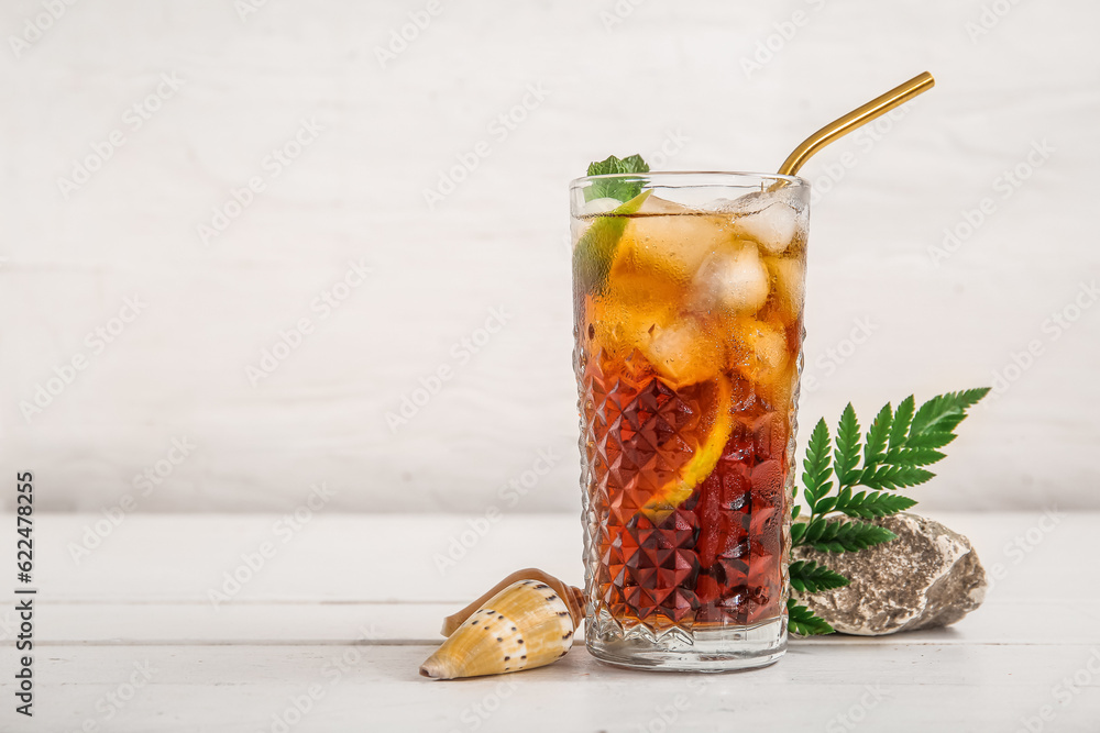 Glass of cold Cuba Libre cocktail, stone and seashells on white wooden table