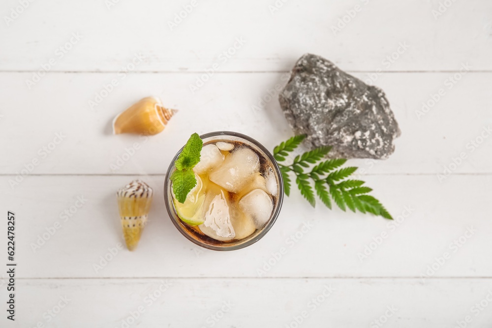 Glass of cold Cuba Libre cocktail, stone and seashells on white wooden background