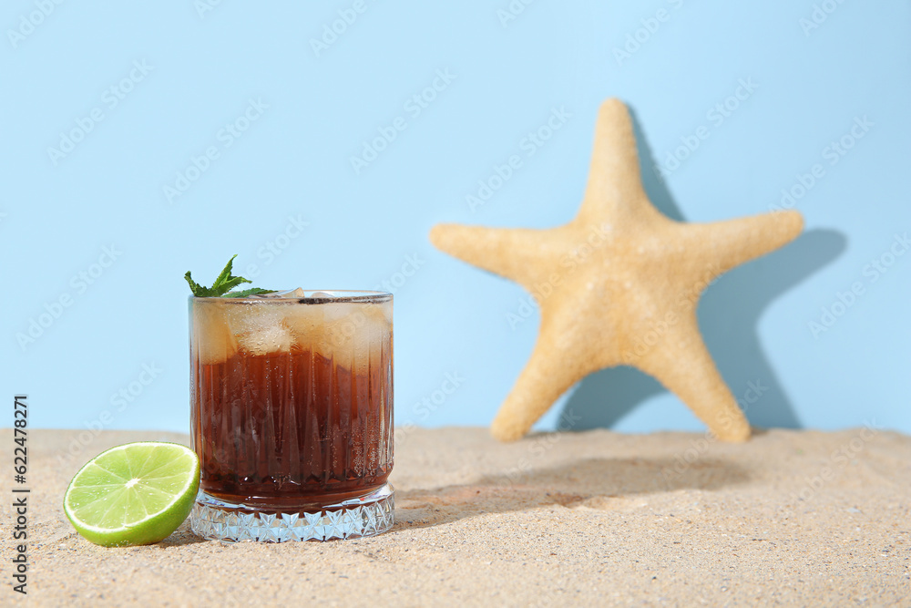 Glass of cold Cuba Libre cocktail and starfish on sand near blue background