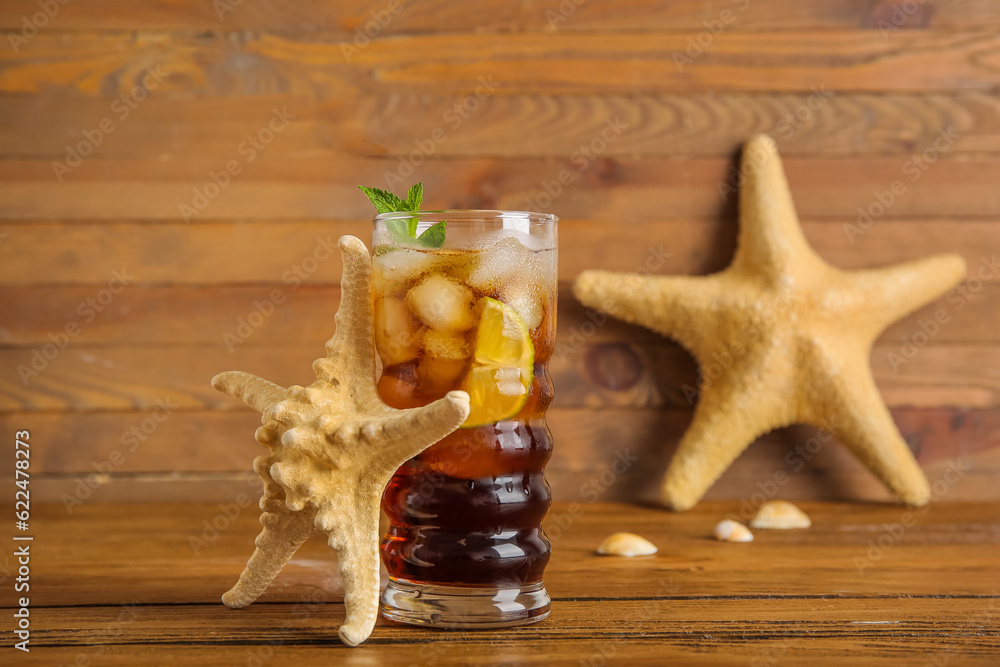 Glass of cold Cuba Libre cocktail, starfishes and seashells on wooden background