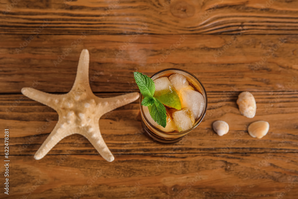 Glass of cold Cuba Libre cocktail, starfish and seashells on wooden background