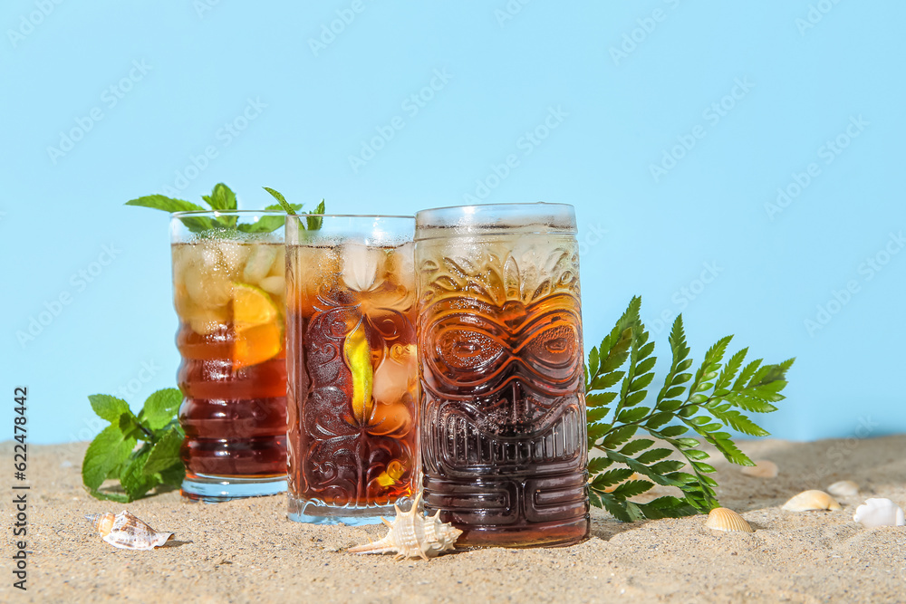 Glasses of cold Cuba Libre cocktail and seashells on sand near blue background