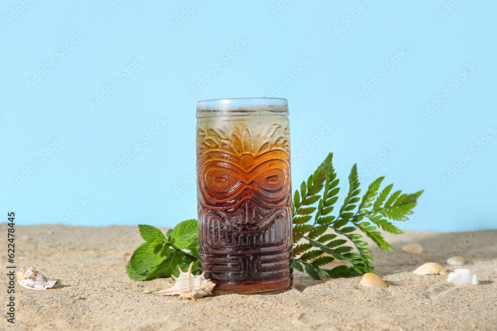 Glass of cold Cuba Libre cocktail and seashells on sand near blue background