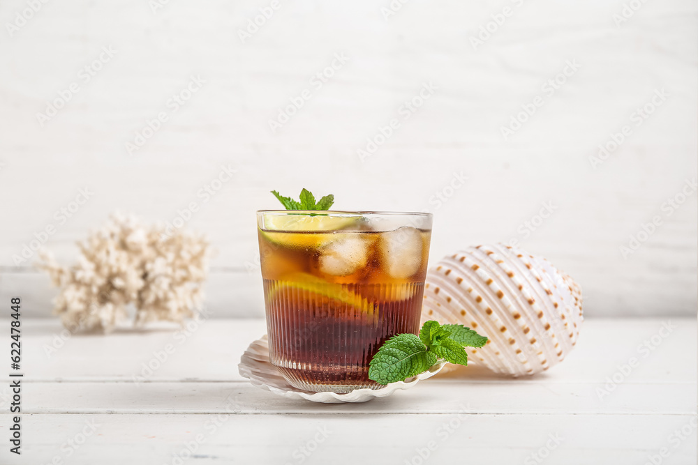 Glass of cold Cuba Libre cocktail, coral and seashells on white wooden table