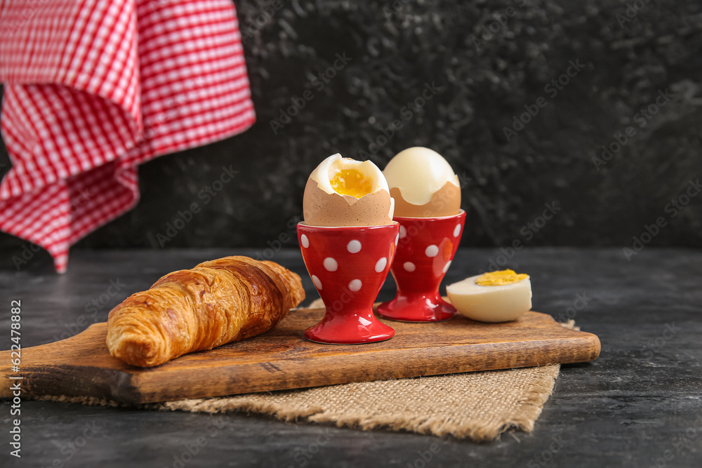 Holders with boiled chicken eggs and croissant on black background