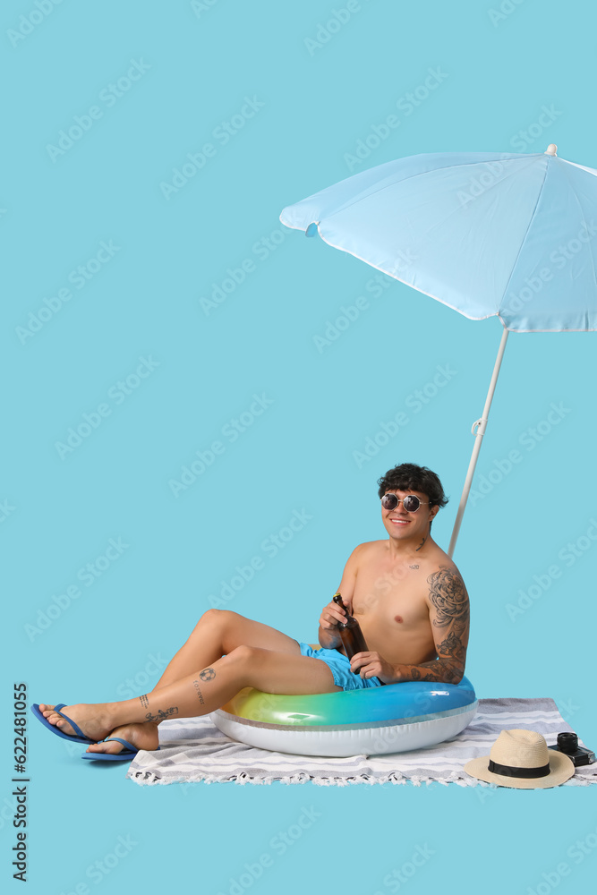 Young man with beer and beach accessories sitting on blue background