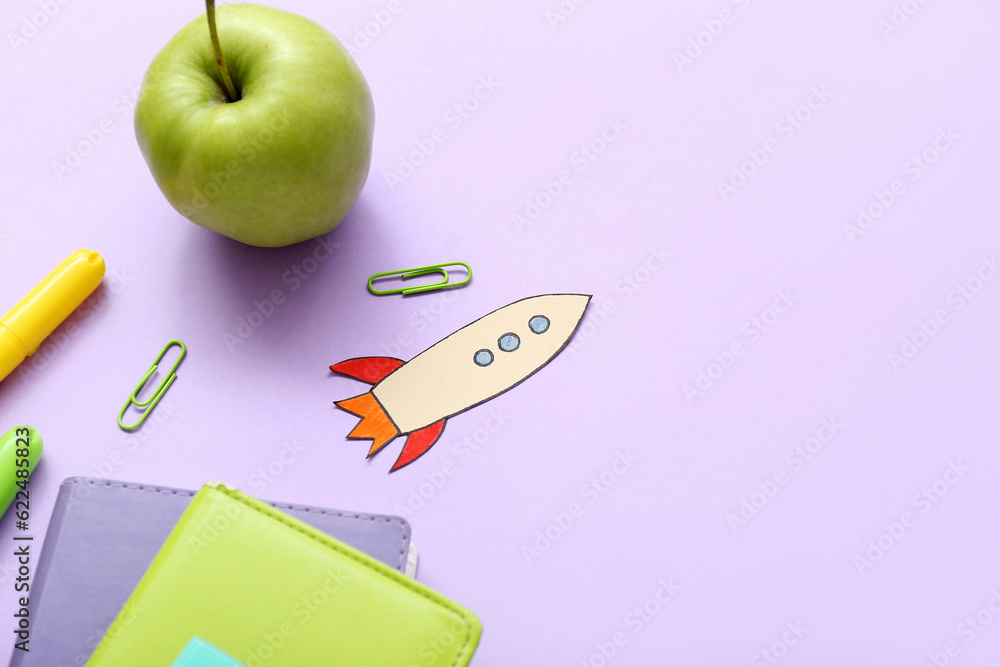 Paper rocket with school stationery and apple on purple background