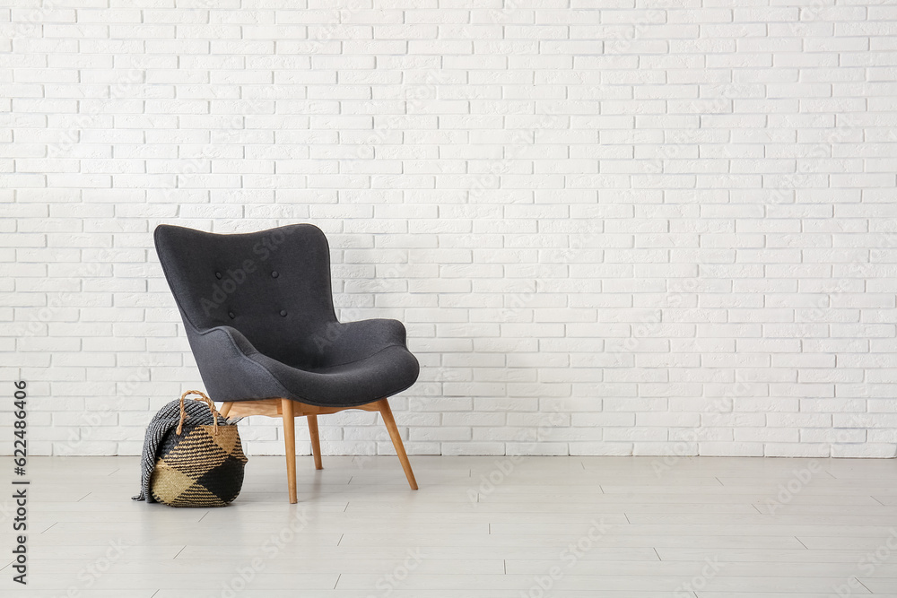 Stylish black armchair with wicker bag and blanket near white brick wall