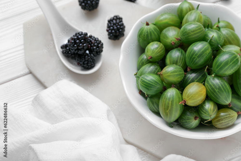 Bowl with fresh green gooseberry and blackberry on light wooden background, closeup