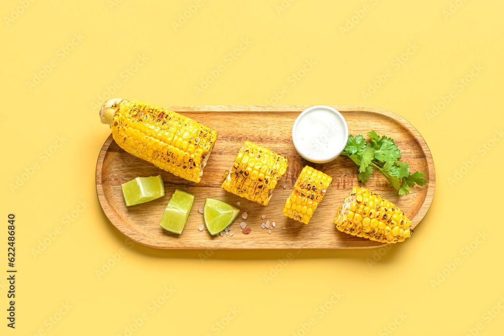 Wooden board with cut tasty grilled corn cob and sauce on yellow background