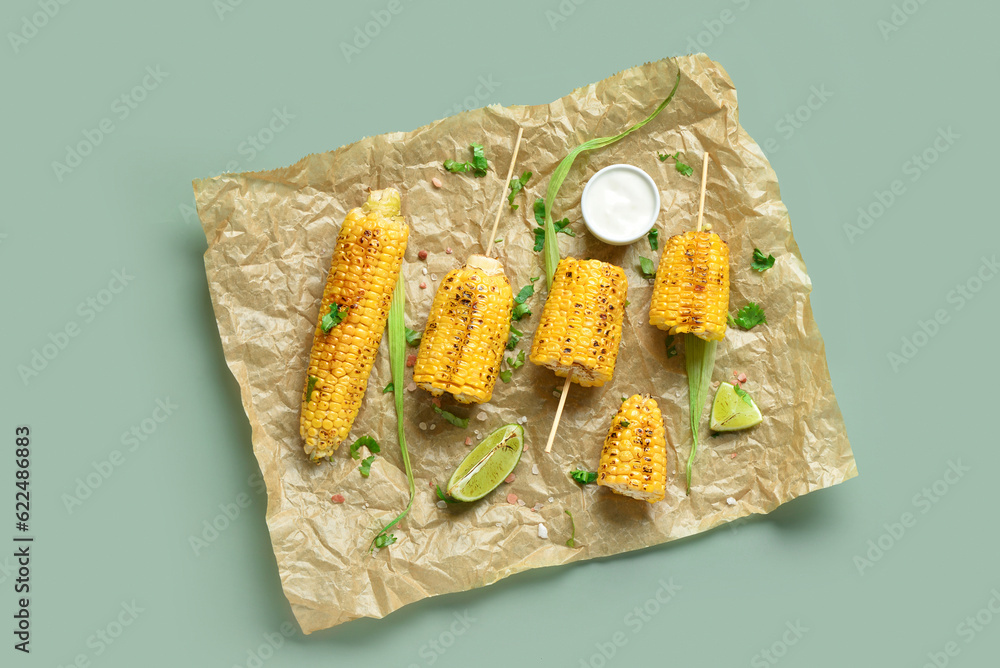 Baking paper with tasty grilled corn cob skewers and sauce on green background