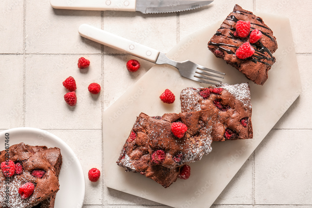 Board and plate with pieces of raspberry chocolate brownie on white tile background