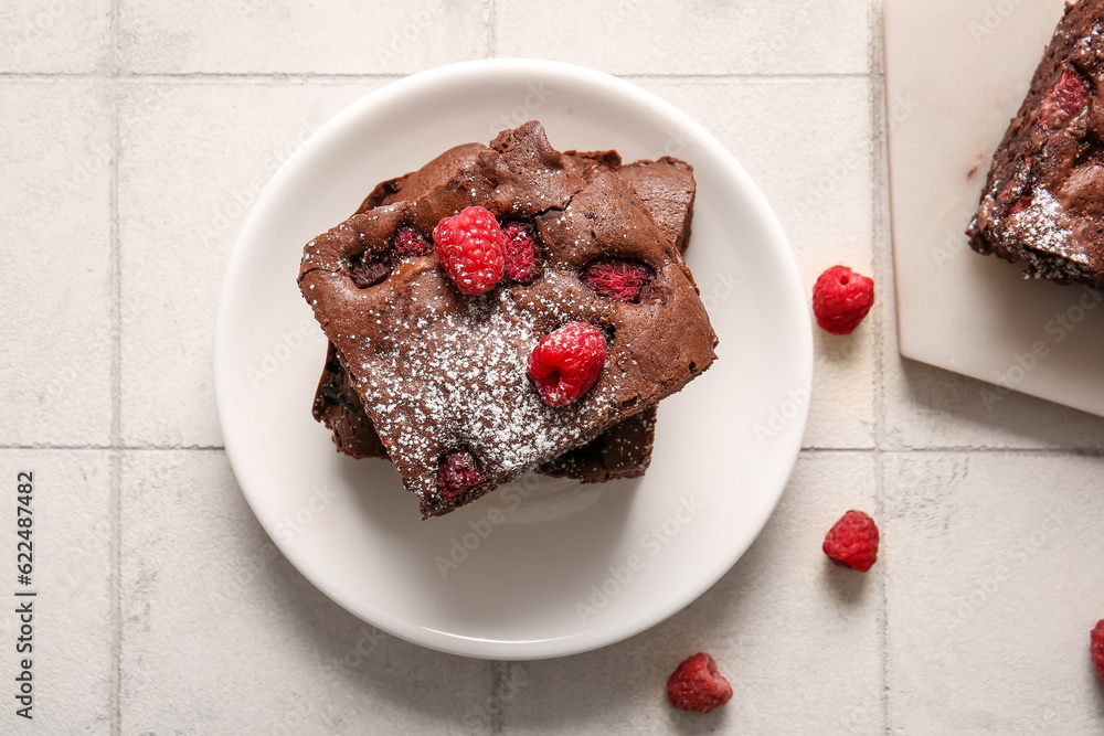 Plate with pieces of raspberry chocolate brownie on white tile background