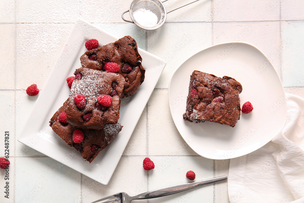 Plates with pieces of raspberry chocolate brownie on white tile background