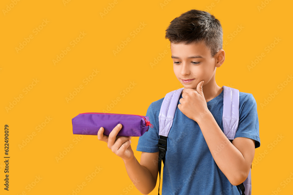 Thoughtful little schoolboy with pencil case on orange background