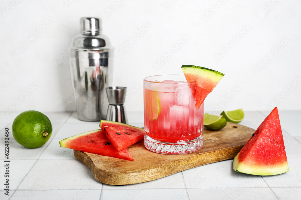 Board with glass of tasty watermelon cocktail, shaker, jigger and lime on white tile table