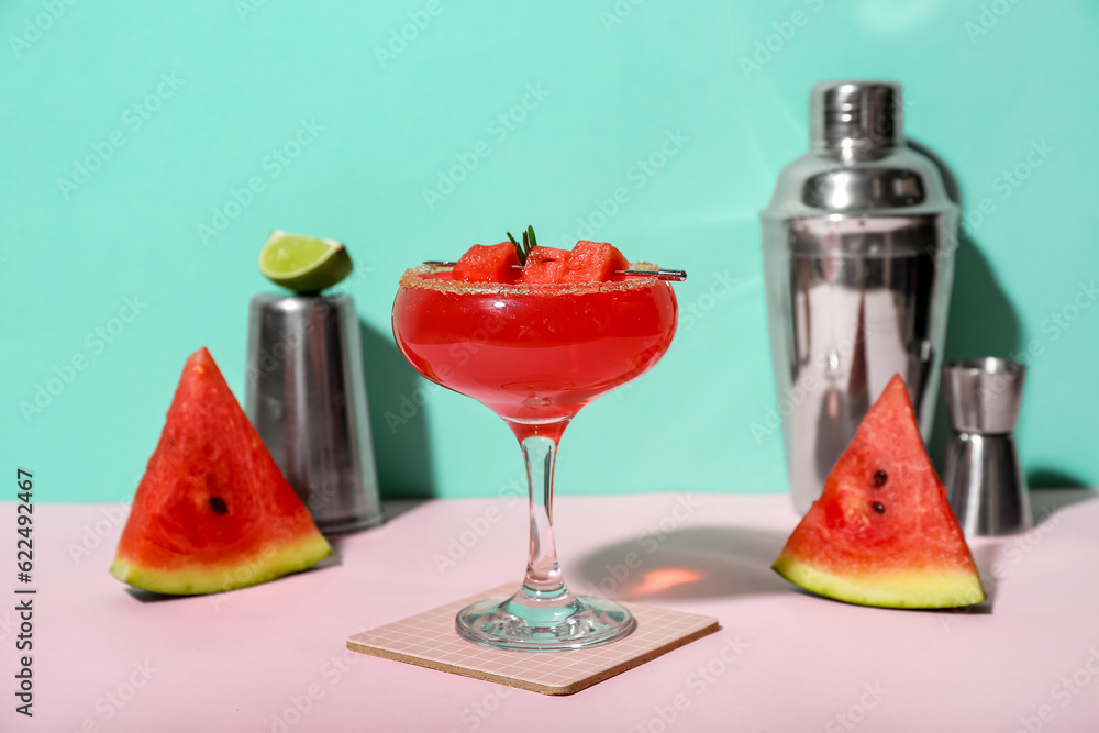 Glass of tasty watermelon cocktail, shakers and jigger on pink table near turquoise wall