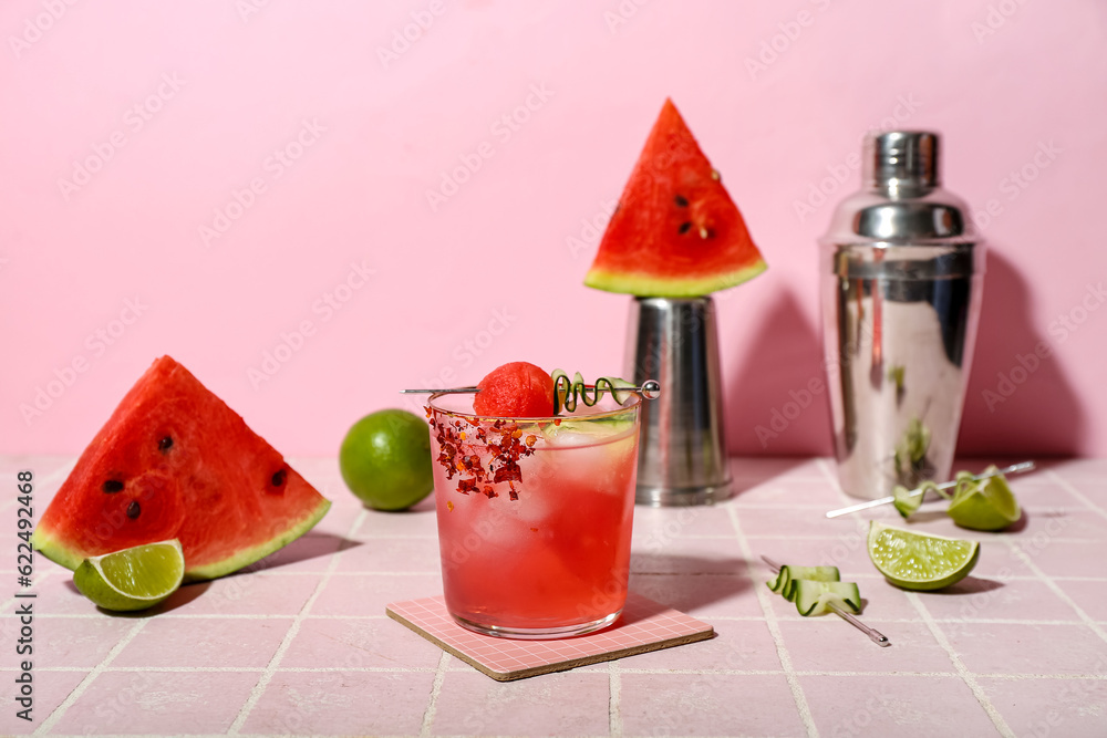 Glass of tasty watermelon cocktail with shakers and lime on tile table near pink wall