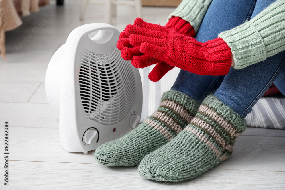 Woman warming hands in gloves near electric fan heater at home, closeup