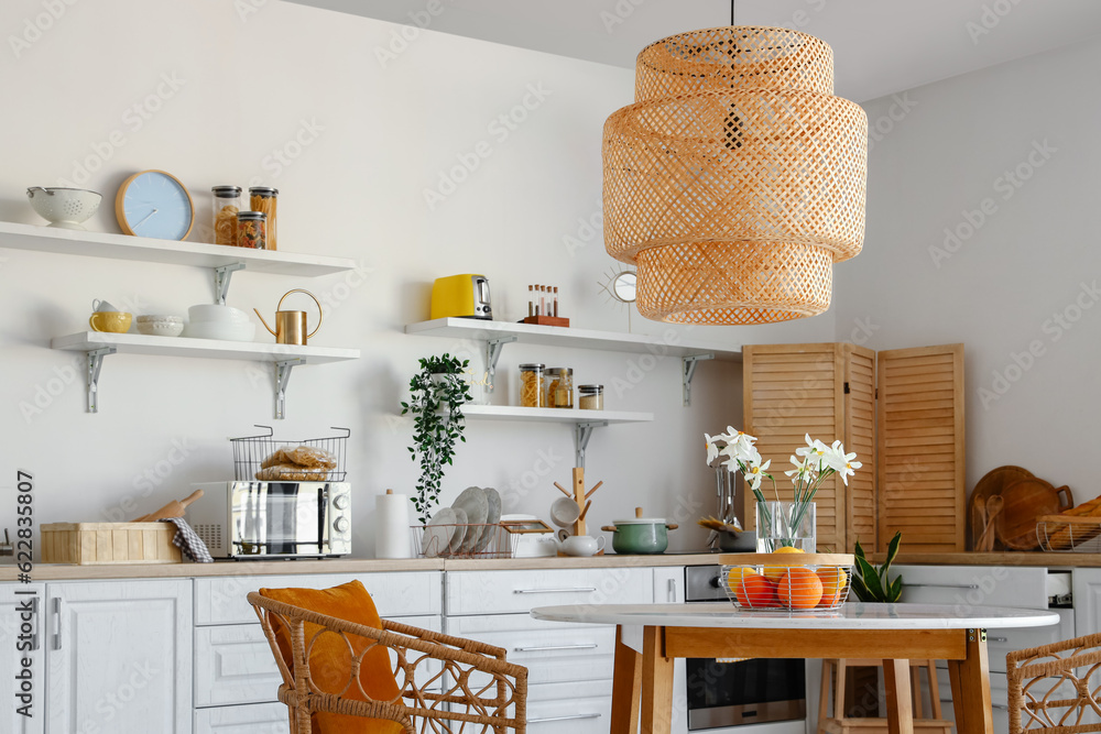 Interior of light kitchen with stylish counters, shelves, table and lamp