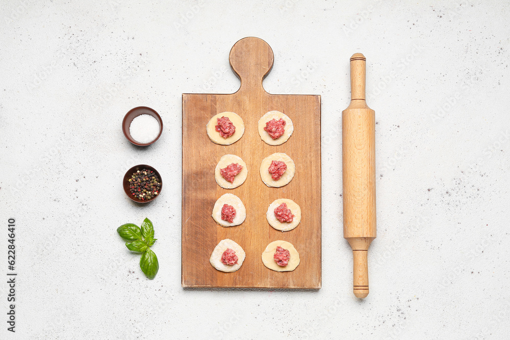 Wooden board of raw dough with minced meat and ingredients for preparing dumplings on light backgrou