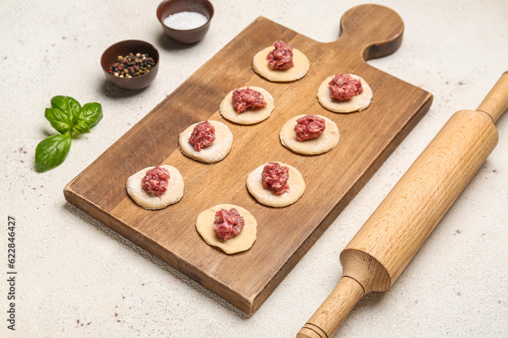 Wooden board of raw dough with minced meat and ingredients for preparing dumplings on light backgrou