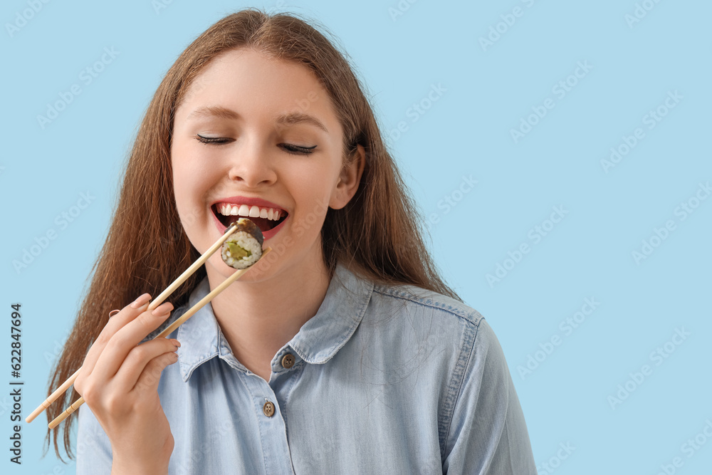 Young woman eating tasty sushi roll on blue background, closeup