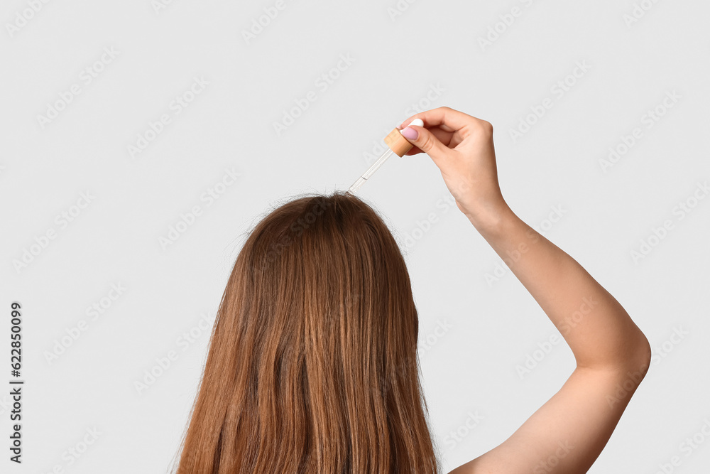 Young woman using cosmetic oil for hair treatment on light background, back view