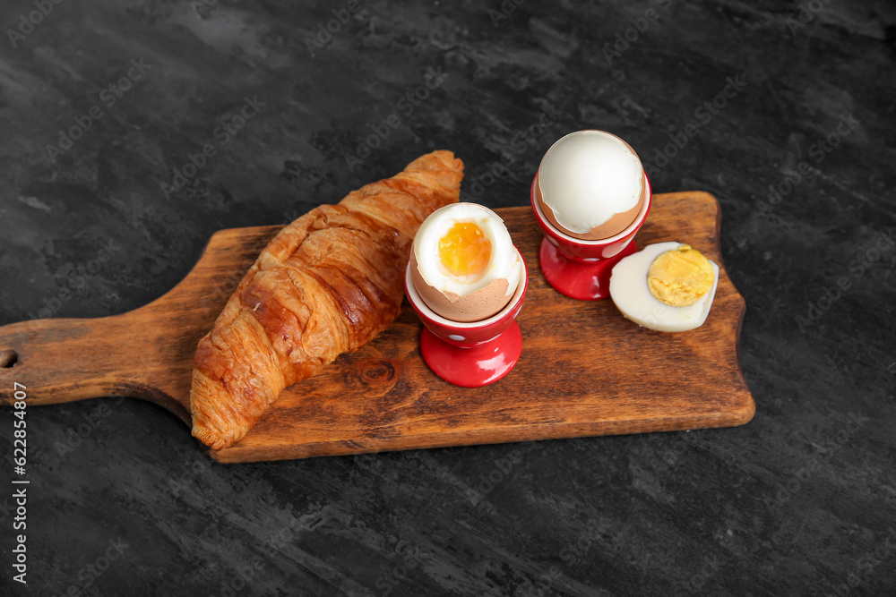 Holders with boiled chicken eggs and croissant on black background