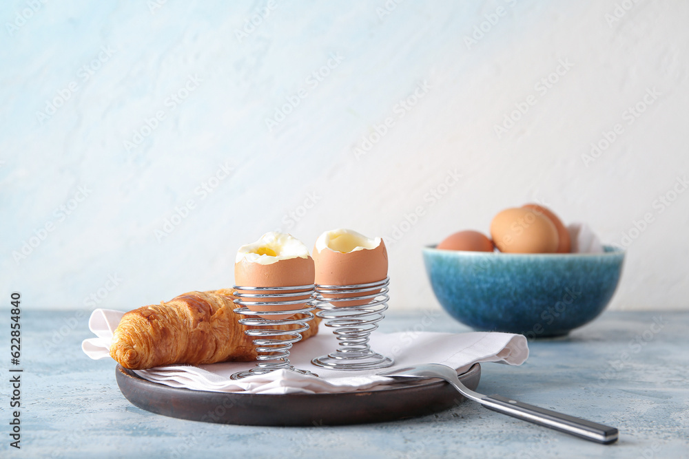 Holders with boiled chicken eggs and croissant on blue table