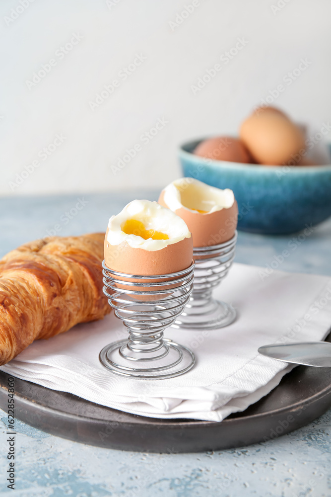 Holders with boiled chicken eggs and croissant on blue table