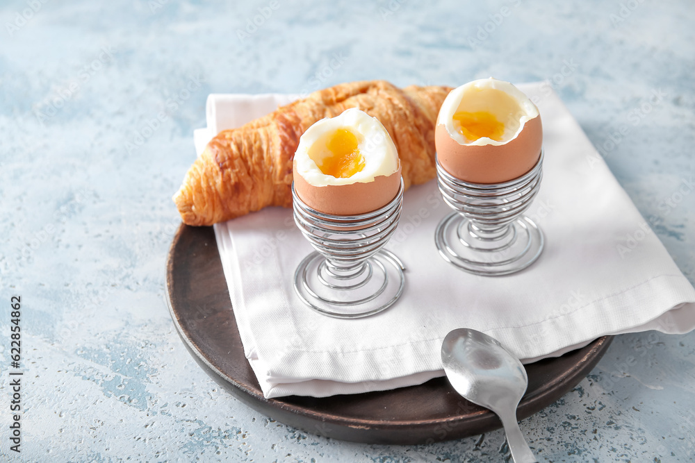 Holders with boiled chicken eggs and croissant on blue table