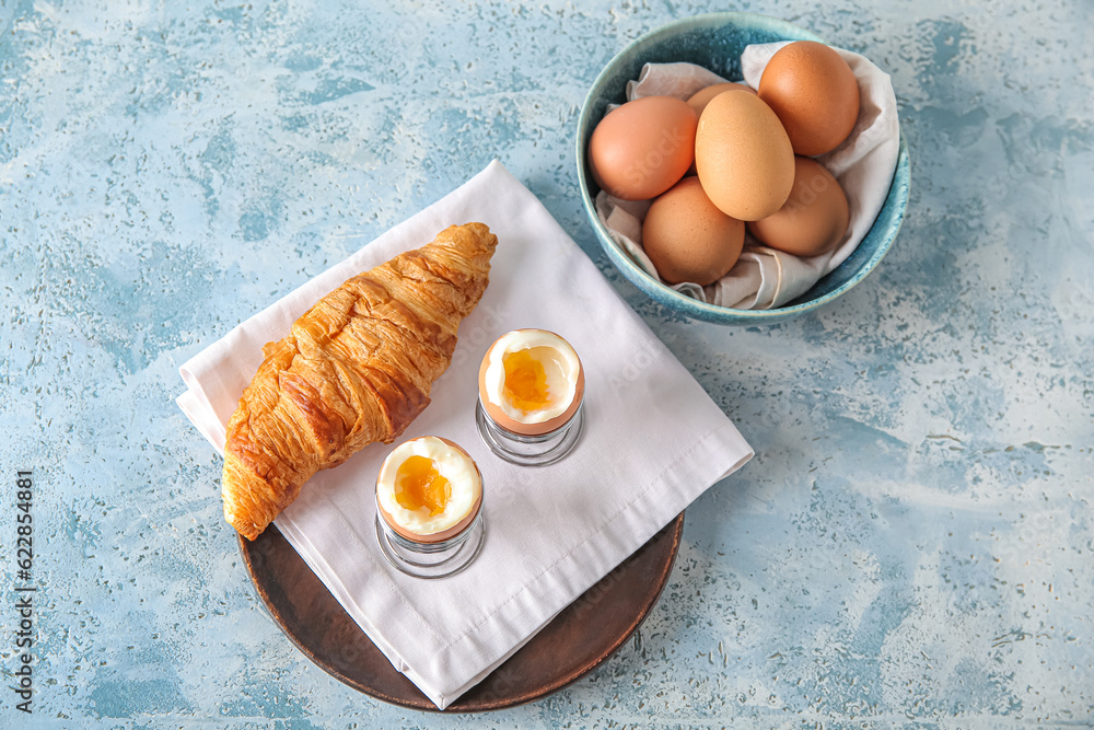 Holders with boiled chicken eggs and croissant on blue table