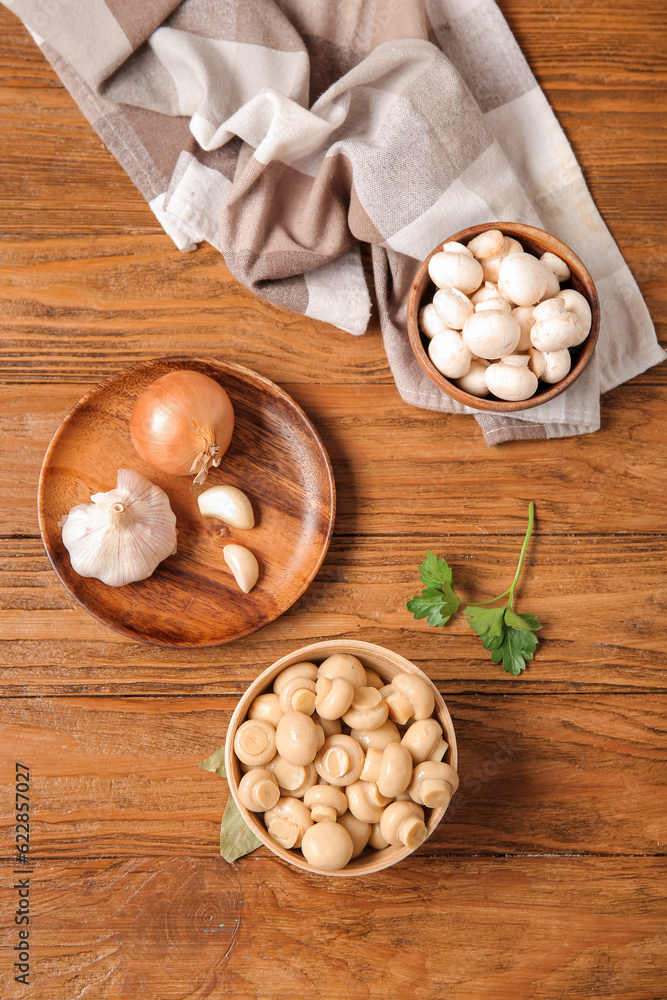 Bowls of canned and fresh mushrooms with ingredients for preservation on wooden background