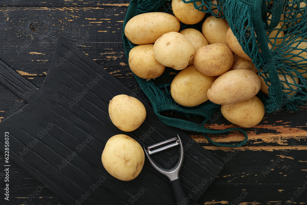 Board and string bag with raw potatoes on black wooden background