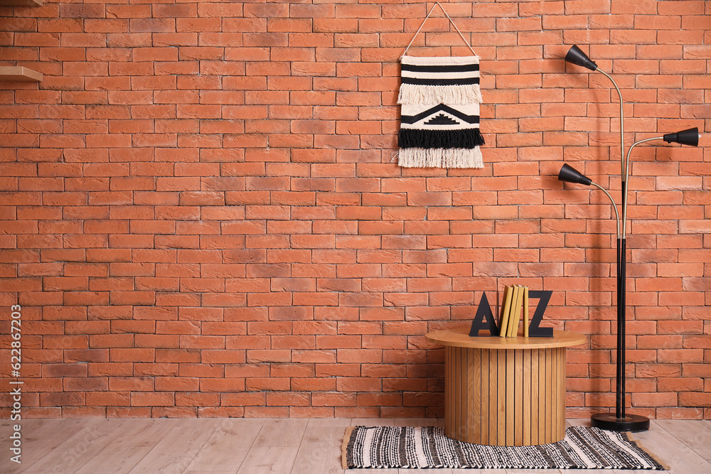 Wooden coffee table with books, standard lamp and macrame near brown brick wall