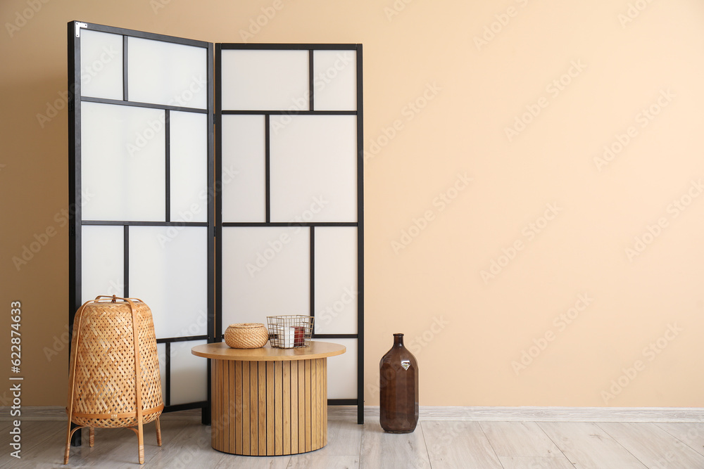 Coffee table with decor, wicker lamp and folding screen near beige wall