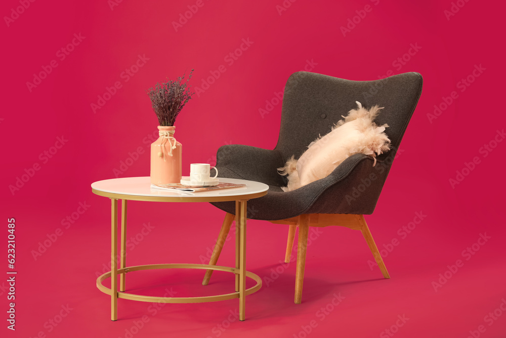 Table with lavender flowers in vase, coffee cup and armchair on pink background