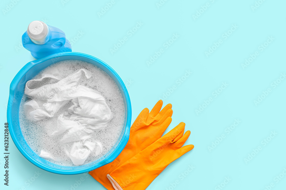 Plastic basin with dirty clothes, water, foam and orange rubber gloves on blue background