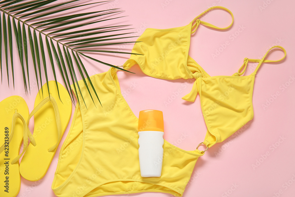 Stylish swimsuit, flip-flops, bottle of sunscreen cream and palm leaf on pink background