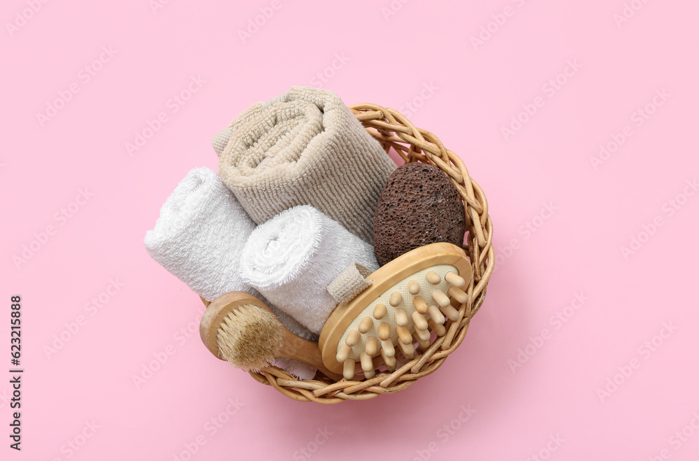 Wicker basket with clean towels, massage brushes and pumice stone on pink background