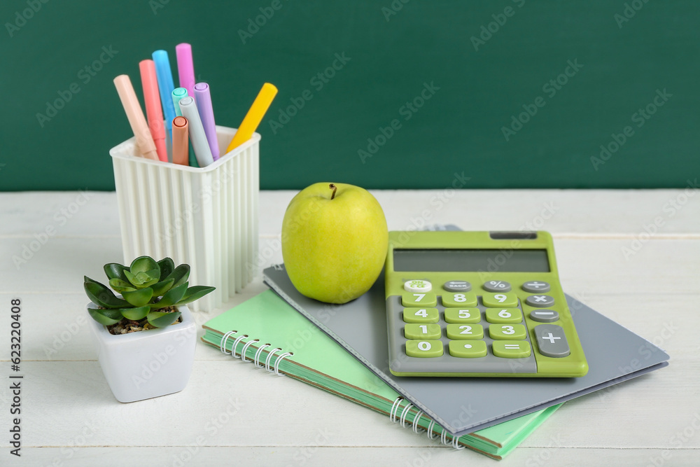 Different stationery with fresh apple and calculator on white table against green chalkboard