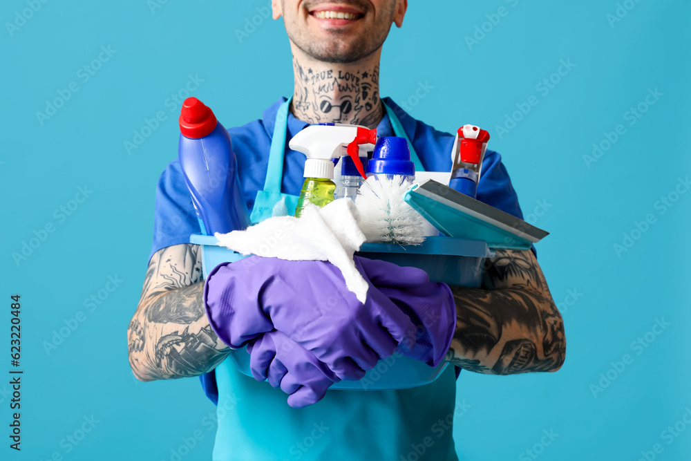 Young tattooed man with bucket of cleaning supplies on blue background, closeup